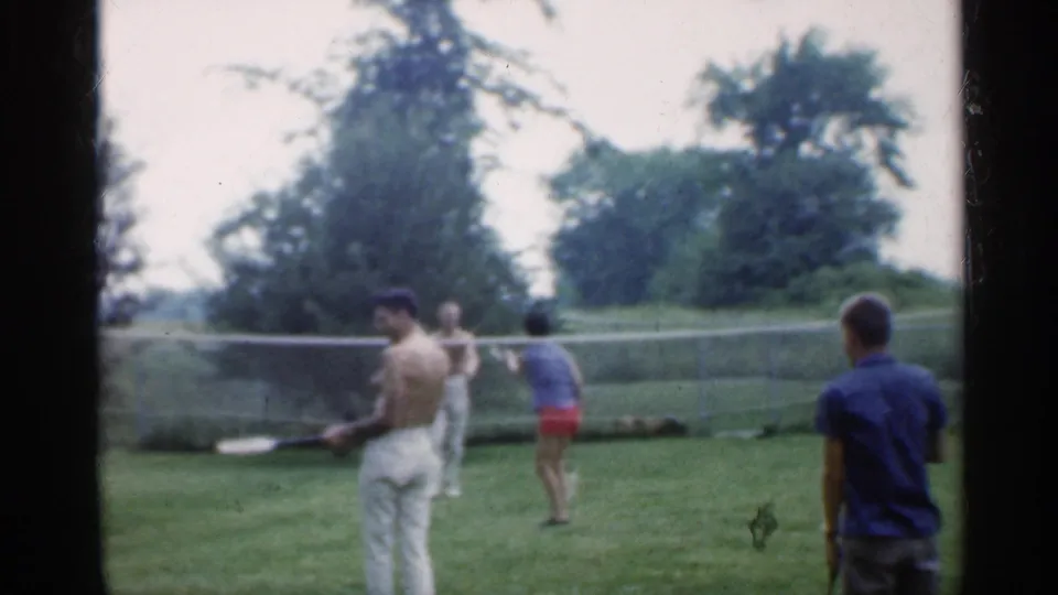Indiana 1959 home-movie frame of adults playing badminton on a grassy lawn beside a chain-link fence.