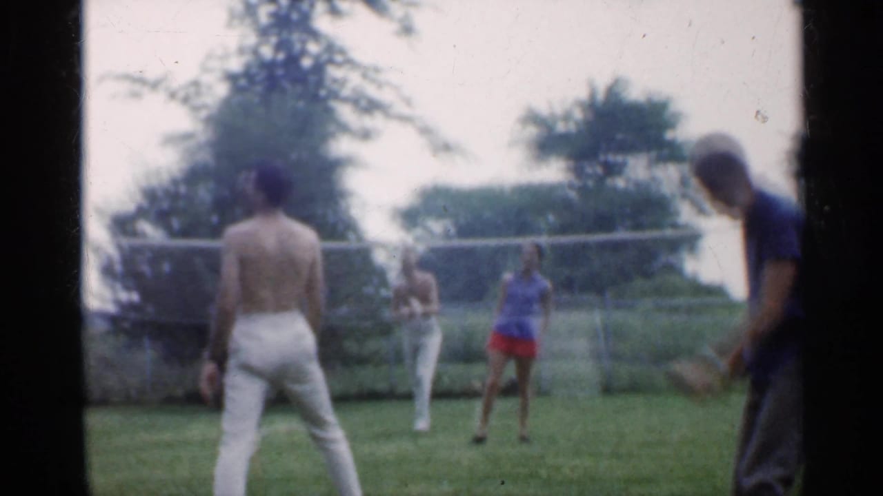 Four adults play badminton on a grassy backyard court with trees and a fence behind them.