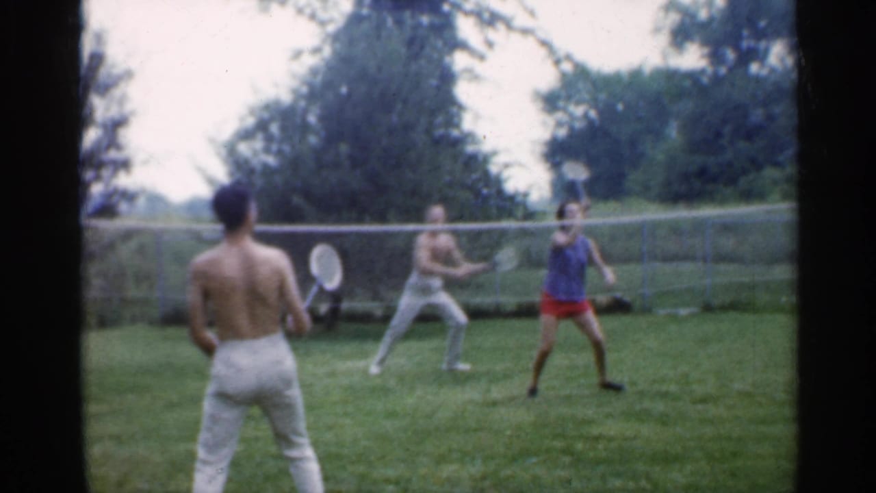 A shirtless man faces three other adults during a backyard badminton rally.