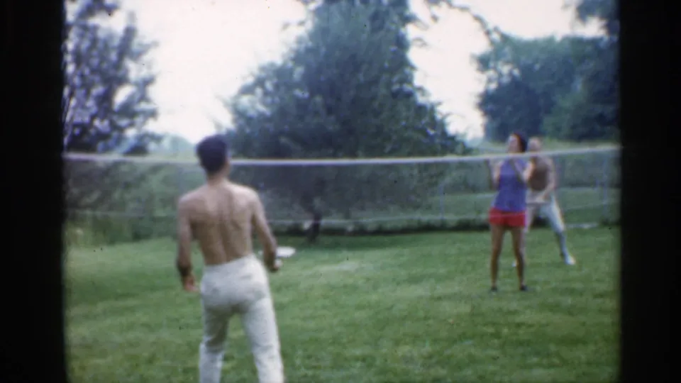 1959 Indiana home-movie still with a shirtless player in the foreground and two adults across a backyard badminton net.