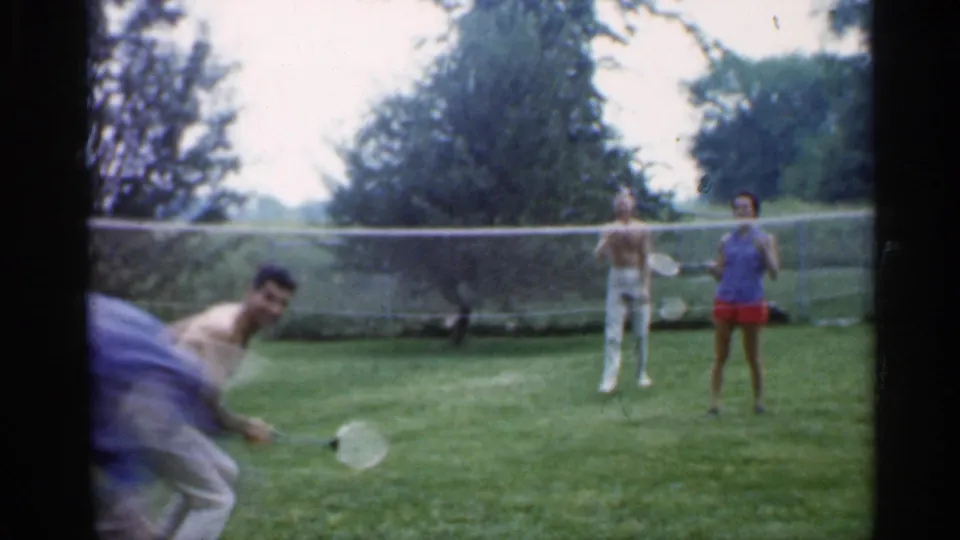 1959 Indiana frame showing a low badminton return in a grassy backyard, with two other adults near the net.