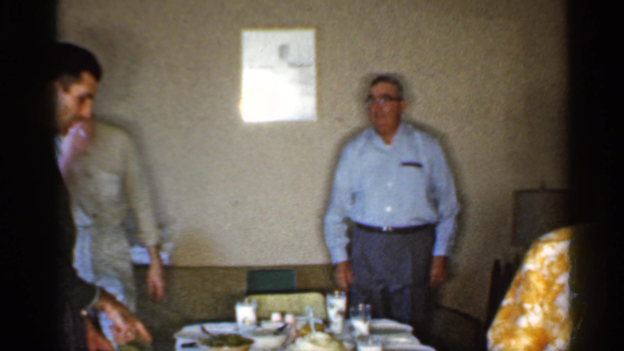 Adult men stand near a set dining table inside a modest home interior.