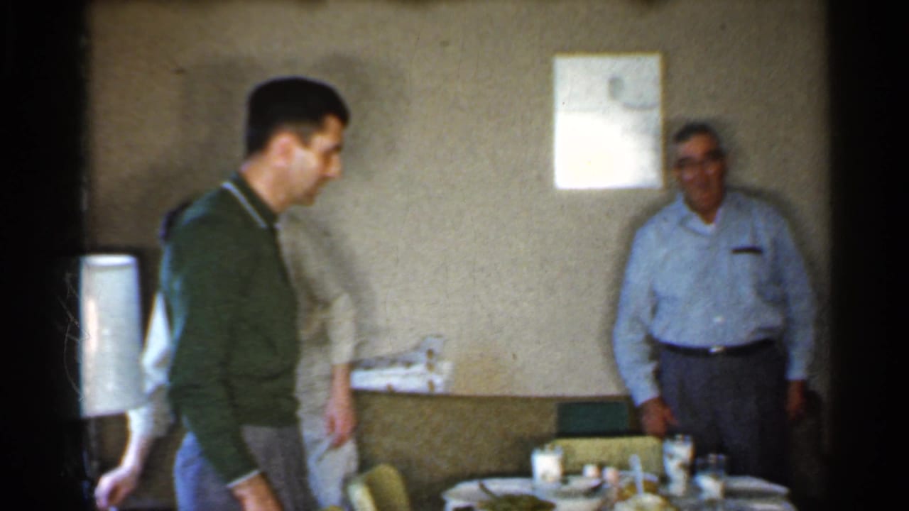 A man in a green sweater moves through a 1959 Indiana home interior as another adult stands near the dining table and wall.