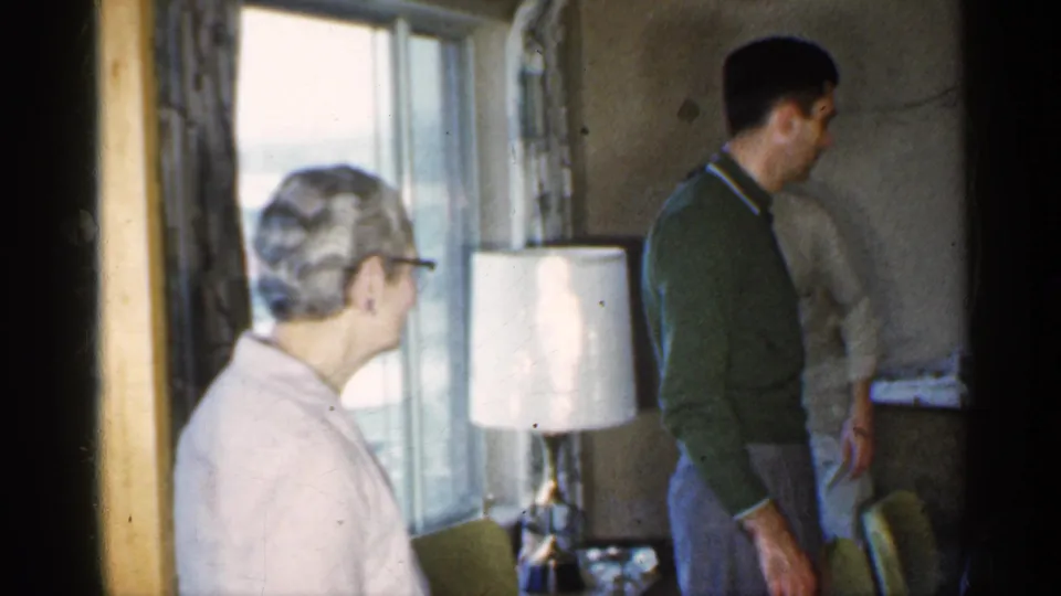 An older woman in a pink cardigan is beside a curtained window as a man in a green sweater stands in the Indiana home interior.