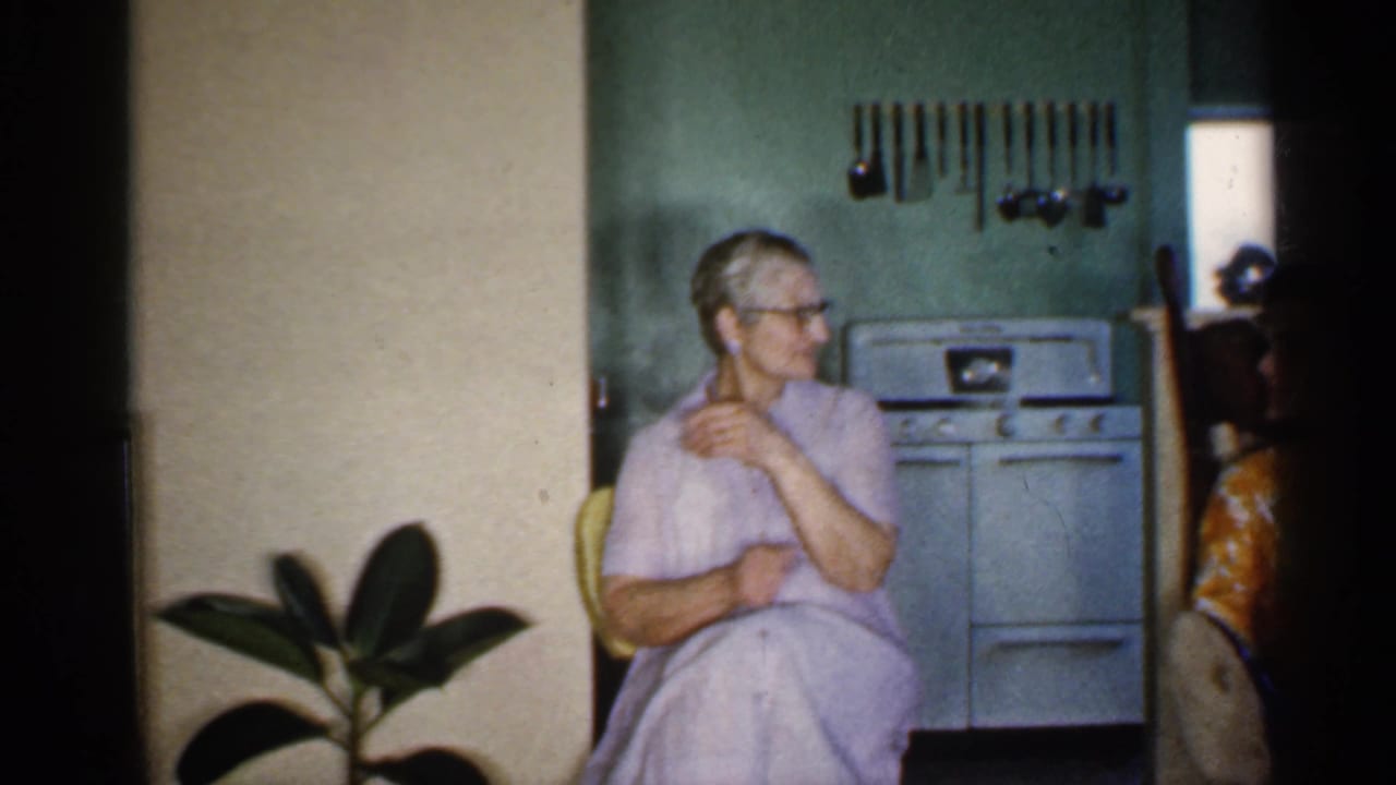 Older woman in a lavender dress sits in a kitchen beside a white stove and potted plant.