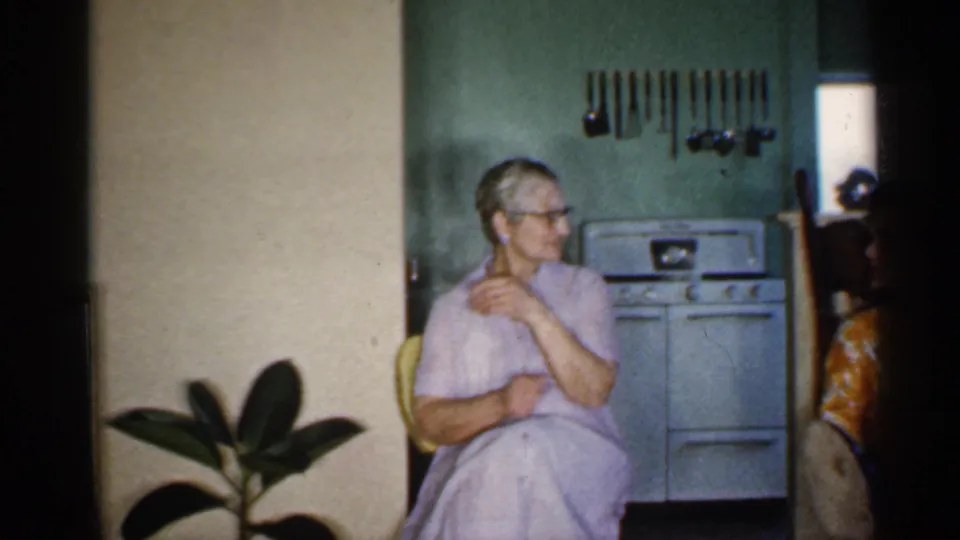 In a 1959 Indiana home-movie frame, an older woman in glasses sits near a white stove, green wall, hanging utensils, and potted plant.
