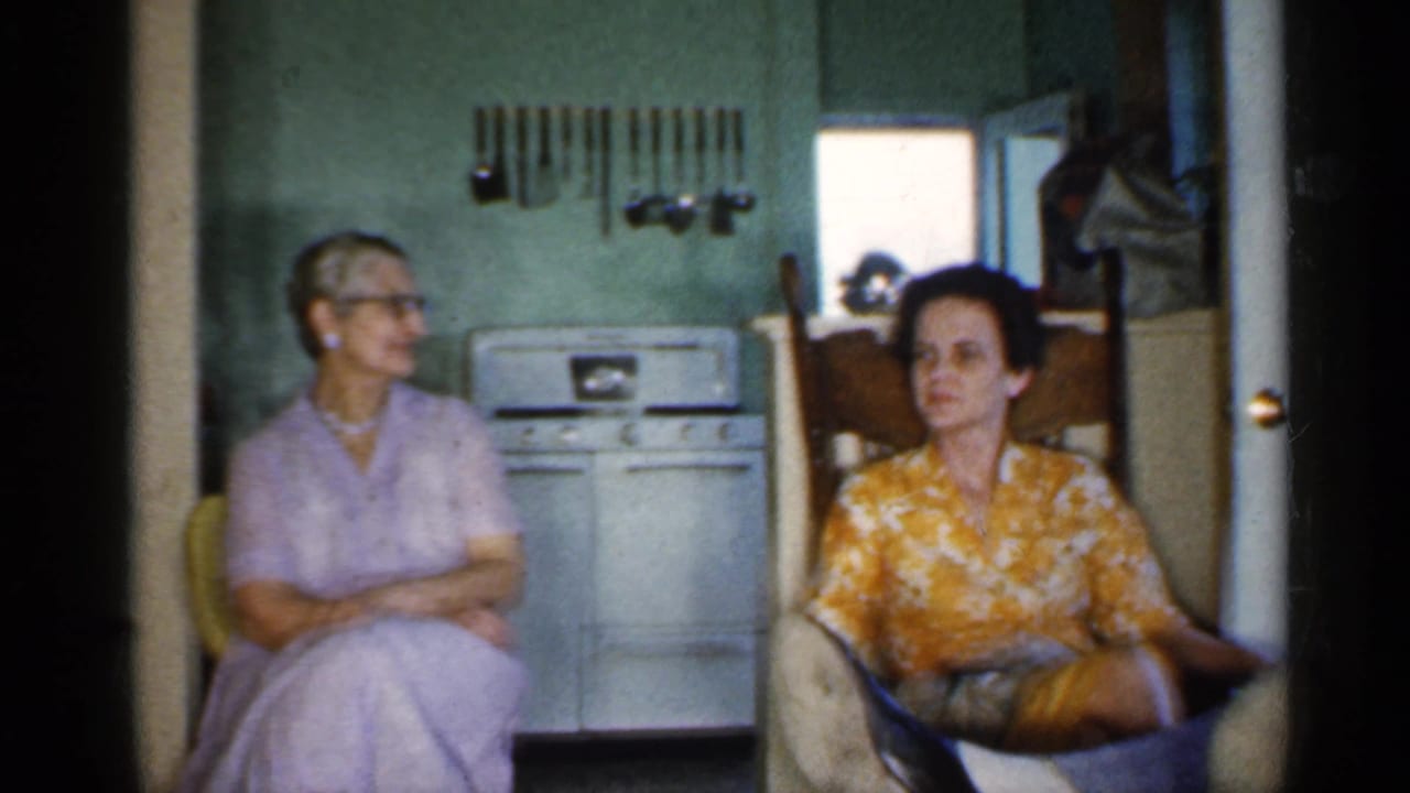 Two women appear seated inside a 1959 Indiana kitchen, one in lavender and one in a yellow floral blouse, with stove and utensils behind them.