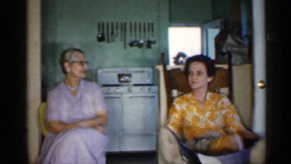 Two women appear seated inside a 1959 Indiana kitchen, one in lavender and one in a yellow floral blouse, with stove and utensils behind them.