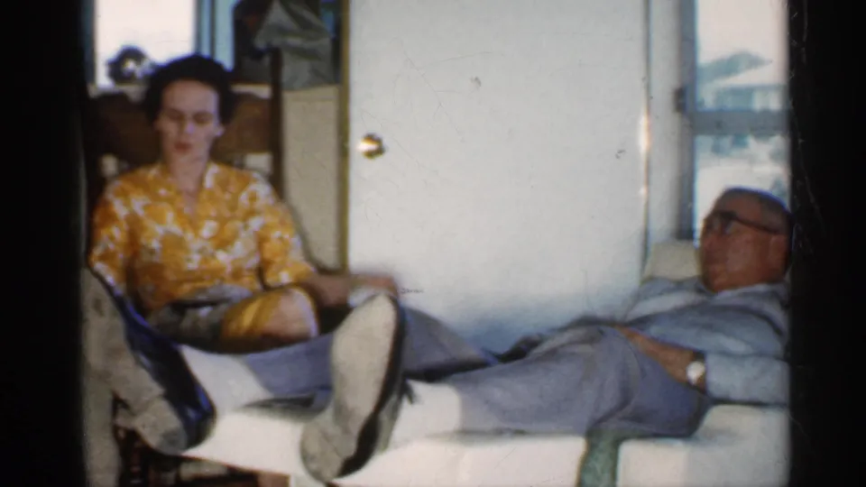 A woman in a yellow floral blouse sits near a man reclining in an upholstered chair inside a private Indiana home-movie interior.