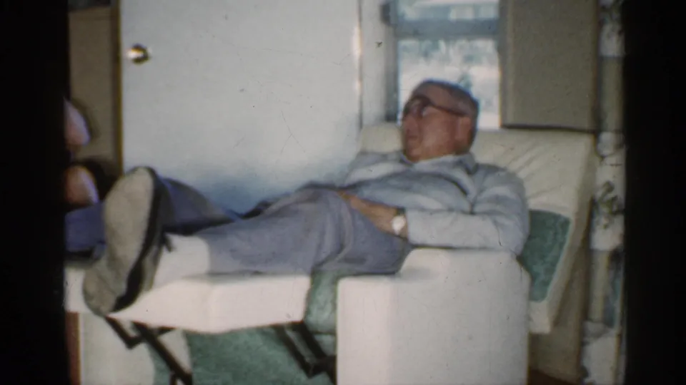A man in glasses reclines in a pale upholstered chair by a window, shown in soft 8mm color within a private 1959 Indiana interior.