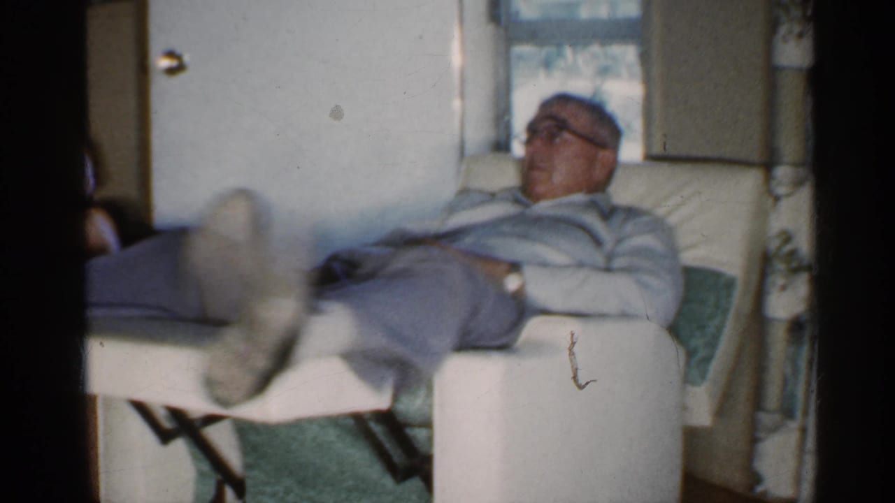 Older man with glasses reclines in a white chair inside a home.