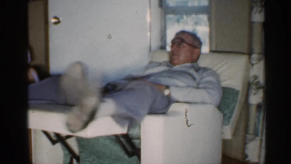 In a 1959 Indiana home interior, an older man wearing glasses reclines in a white lounge chair near a window.