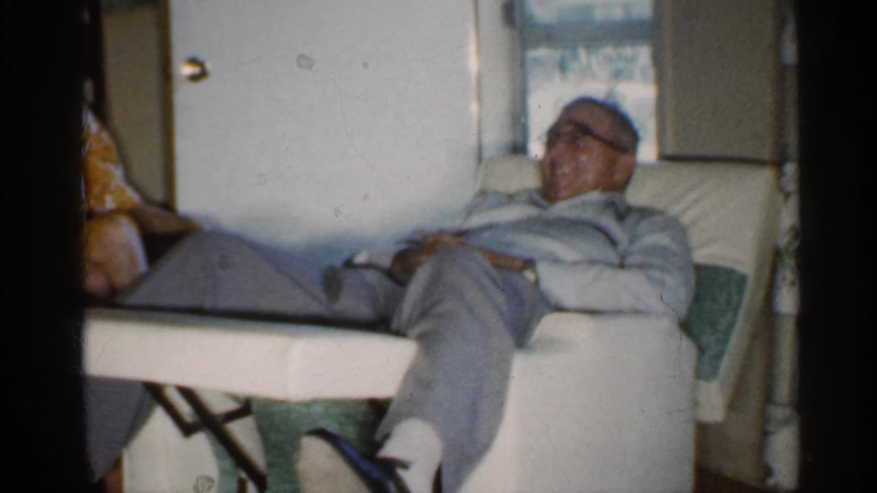 The older man remains reclined in the white chair as a woman in a yellow patterned dress sits partly visible beside him.