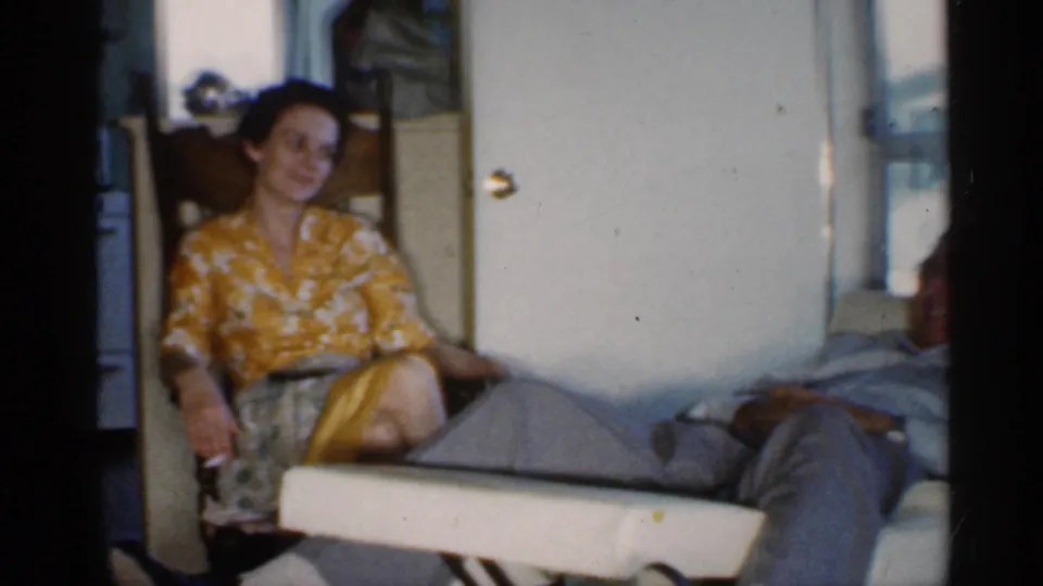 A woman in a yellow patterned dress sits smiling beside the reclining man in a 1959 Indiana home interior.