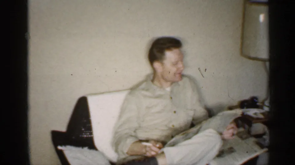 A younger man in a light button shirt sits against a pale wall, looking off to the side in the same indoor setting.