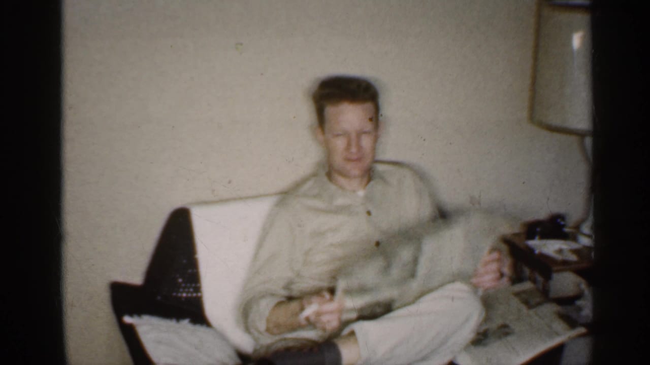 Man seated in a light armchair holding a newspaper beside a lamp and side table.