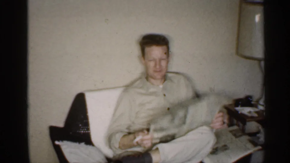 In a 1959 Indiana home interior, a man sits in a light armchair with a newspaper near a lamp and side table.