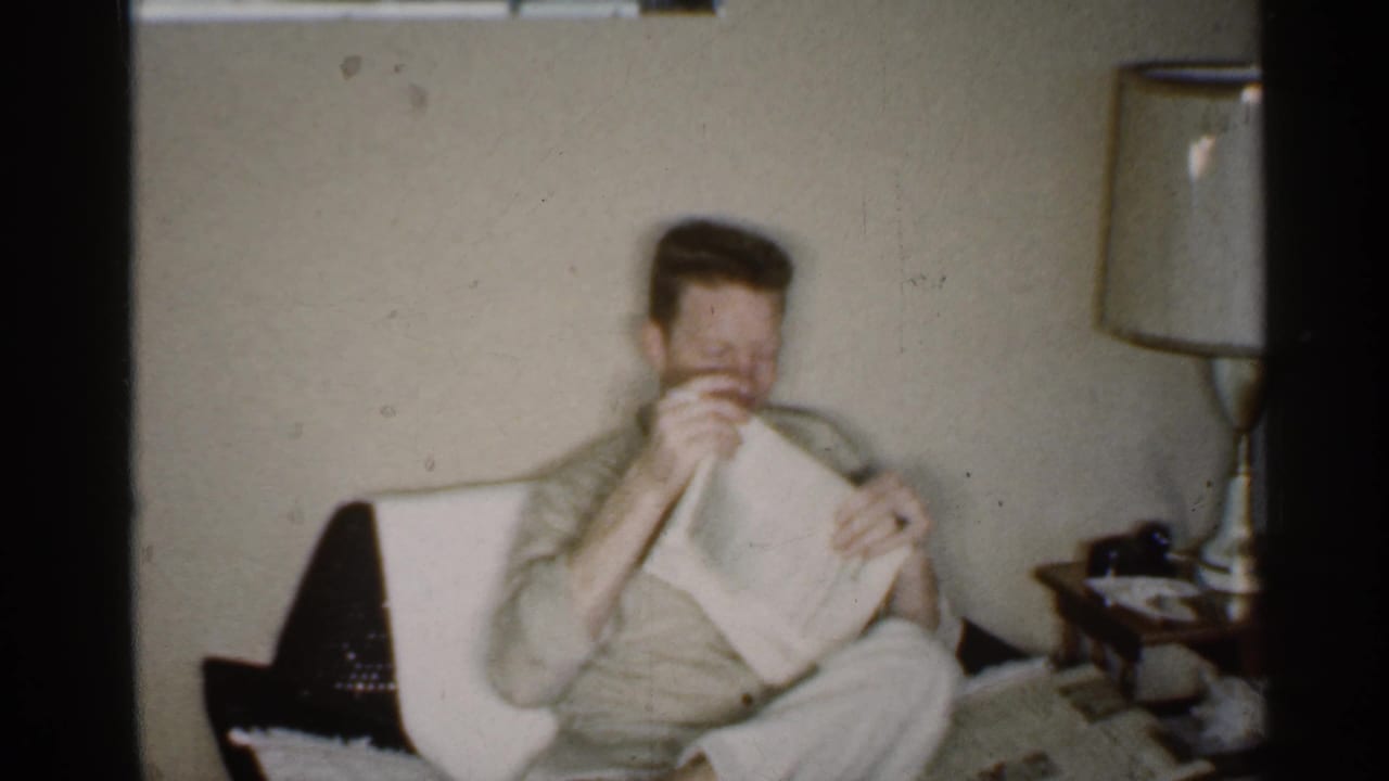 The seated man raises or folds the newspaper in a 1959 Indiana living room with a lamp and side table nearby.
