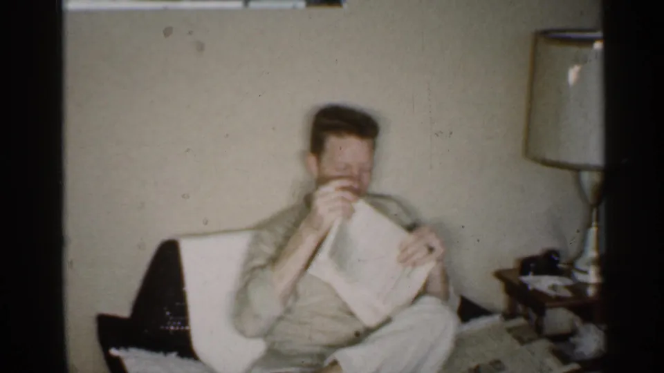 The seated man raises or folds the newspaper in a 1959 Indiana living room with a lamp and side table nearby.