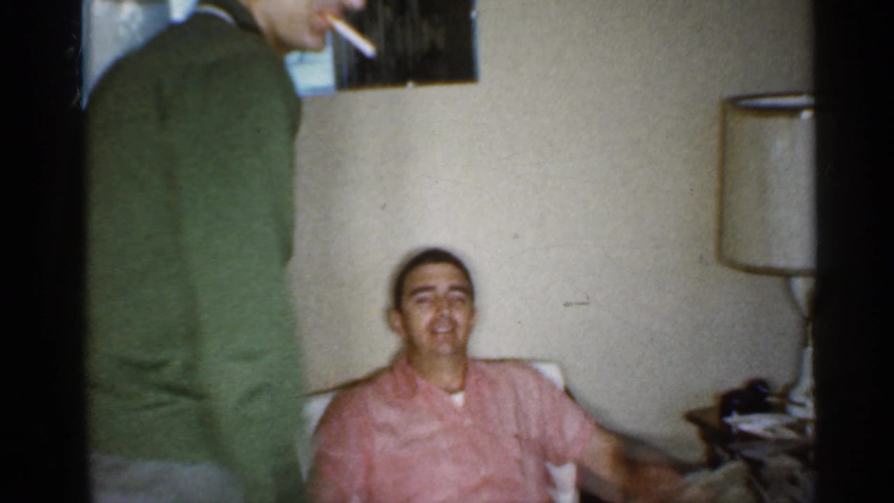 Two adult men in a living room, one standing with a cigarette and one seated in an armchair.