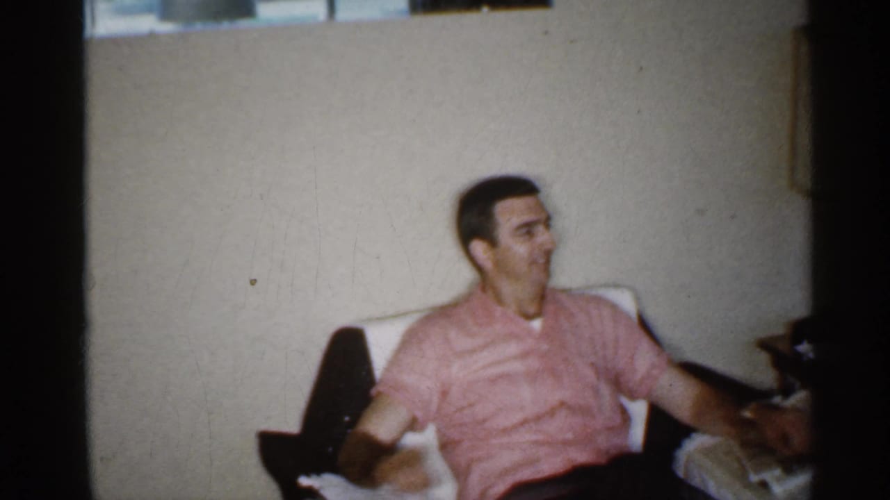 Man in a pink shirt sits in a white armchair beside a table and lamp.