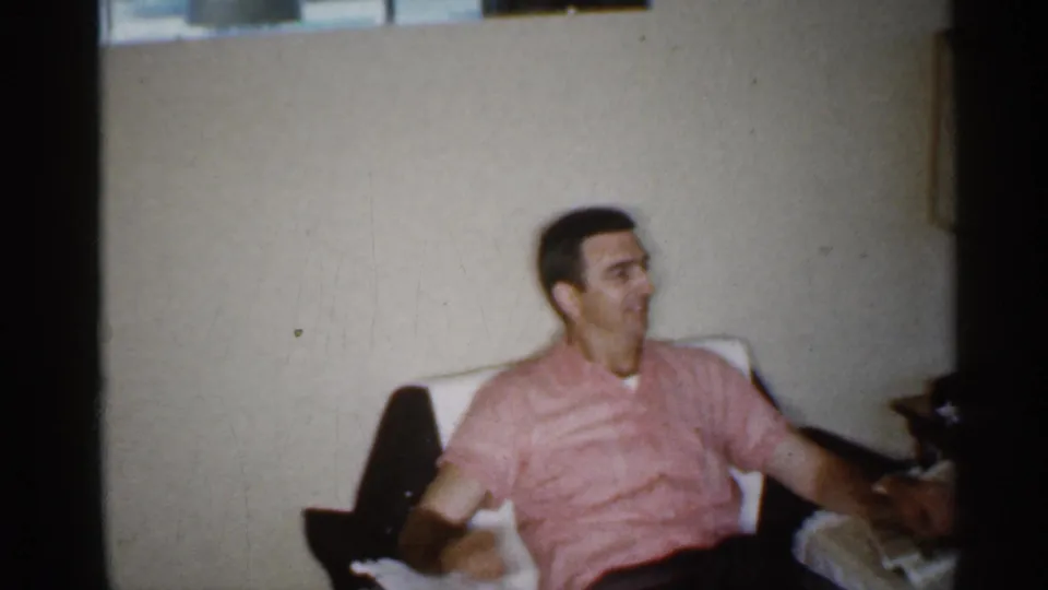 A seated man in a pink striped shirt rests in a white armchair in a 1959 Indiana living room.
