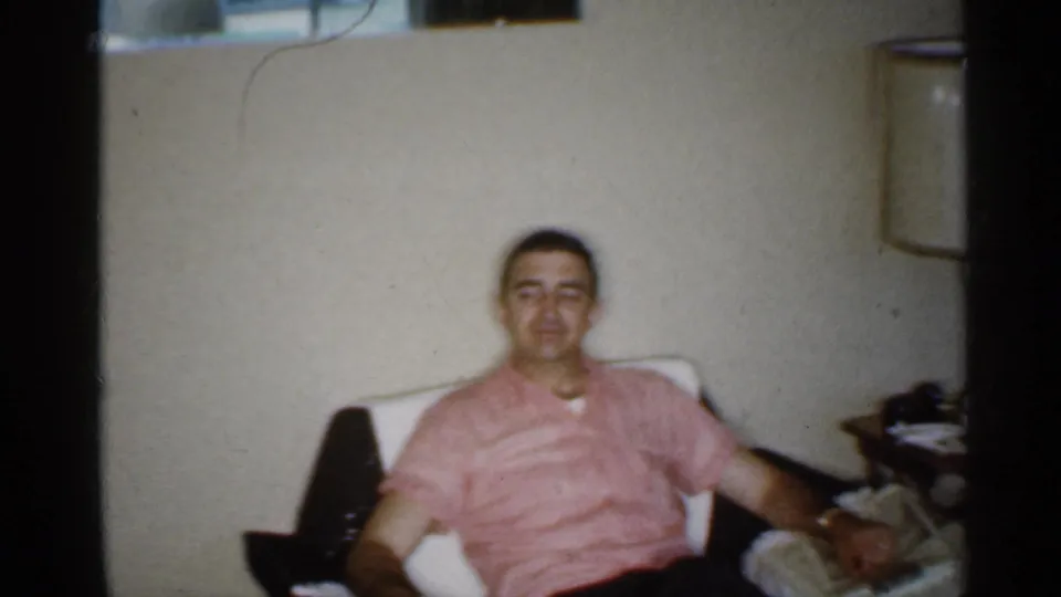 A 1959 Indiana home-movie frame shows a man seated in a white armchair near a lamp and side table.