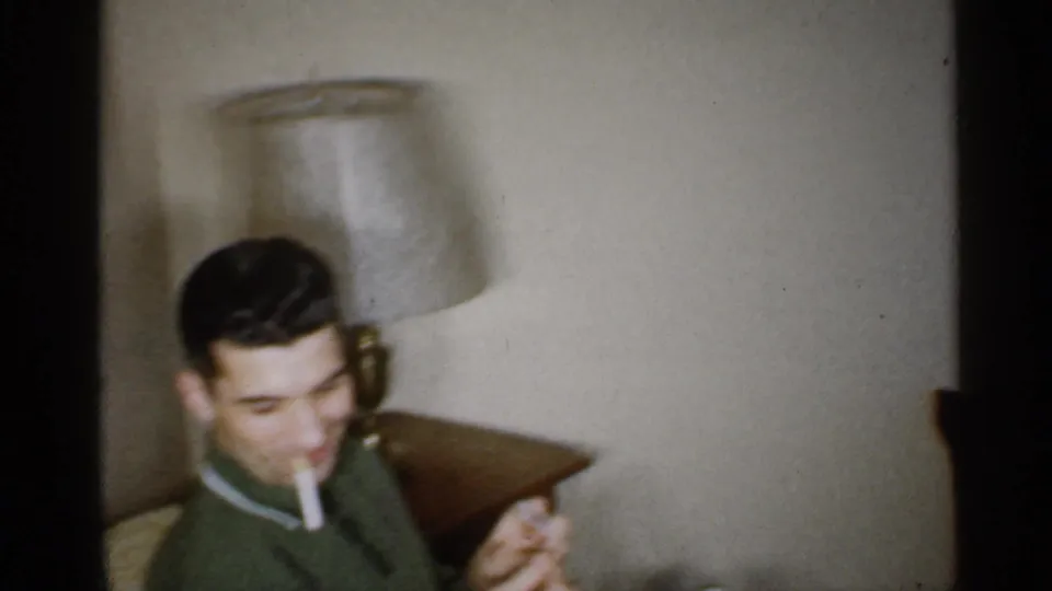A man in a green sweater smokes a cigarette near a table lamp in the Indiana living room footage.