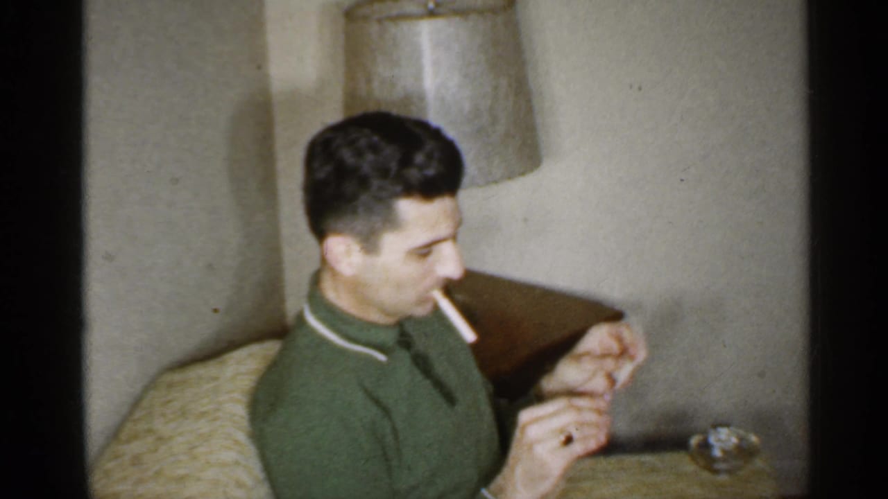 Man in a green shirt sits indoors with a cigarette and raises a match near a couch and lamp.