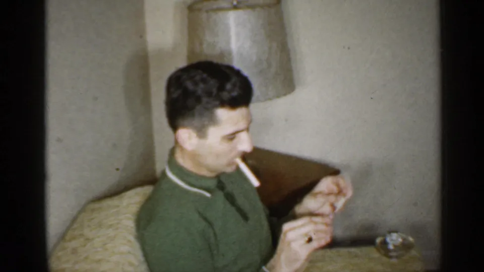 In 1959 Indiana home-movie footage, a man in a green collared shirt sits in a living room while lighting a cigarette beside a lamp.