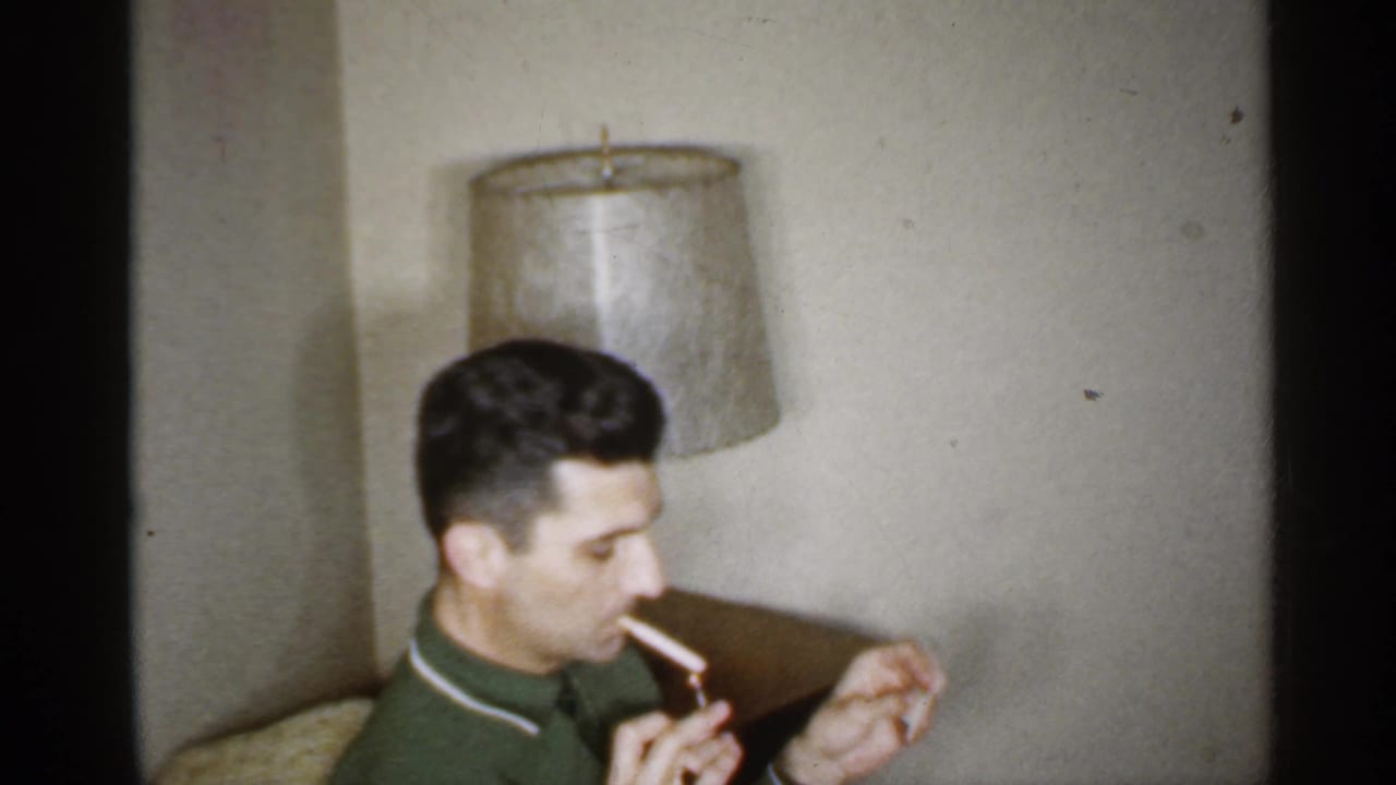 Man sits near a lamp with a cigarette in his mouth and a hand raised in front of him.