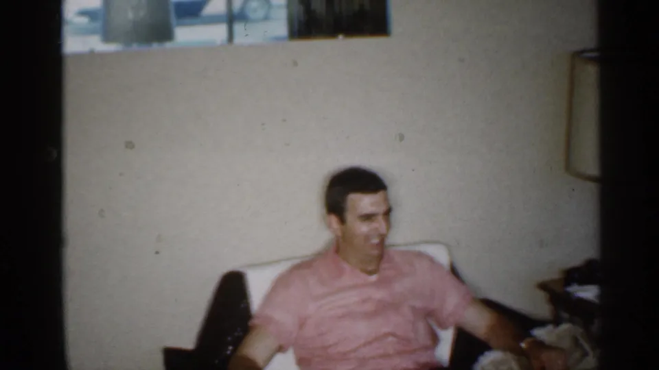 The man in the pink shirt smiles while seated in the Indiana living room, with a chair arm, side furniture, and lamp edge visible.