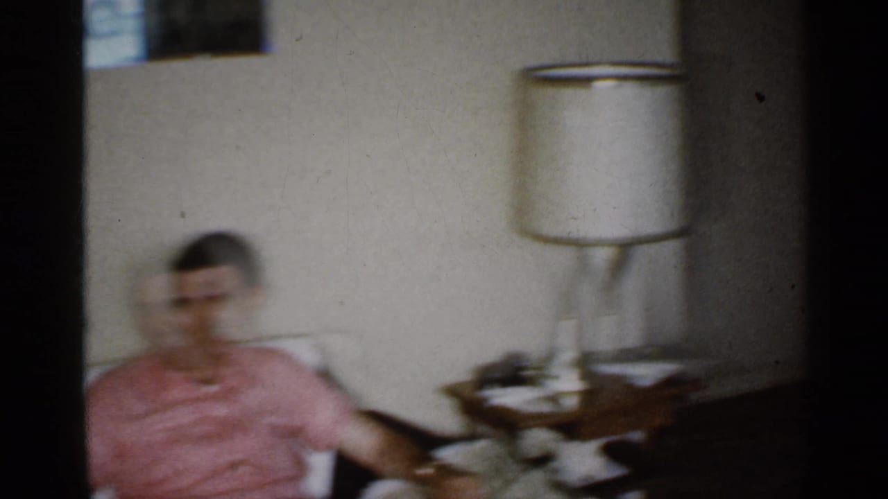 Man in a pink shirt seated beside a table lamp in a living room.