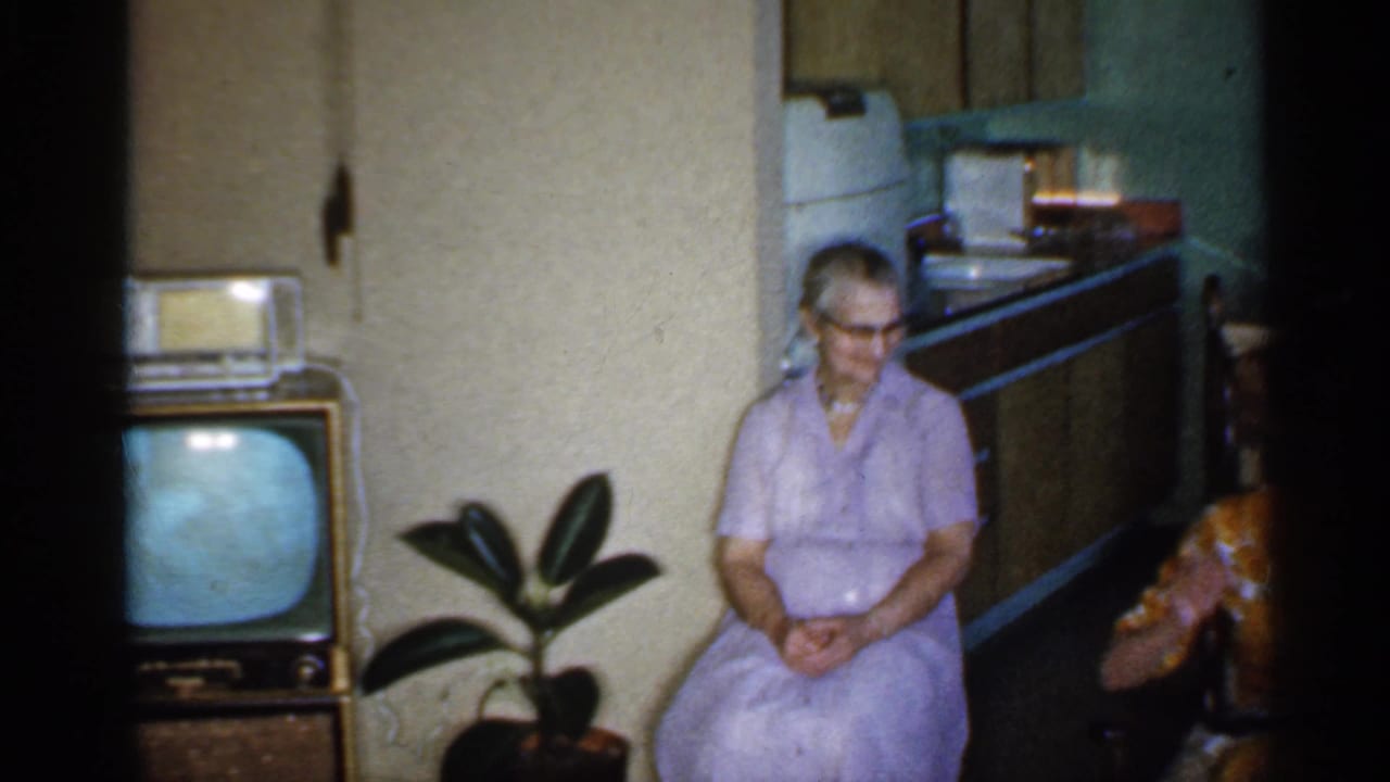 An older woman in a lavender dress sits in a 1959 Indiana living room near a wooden television set, potted plant, and kitchen area.