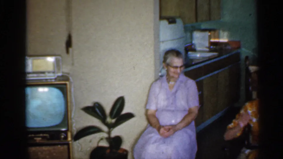 An older woman in a lavender dress sits in a 1959 Indiana living room near a wooden television set, potted plant, and kitchen area.