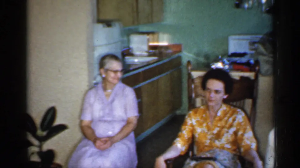 Two women sit near the kitchen side of a 1959 Indiana living room, one in lavender and one in a yellow floral top.
