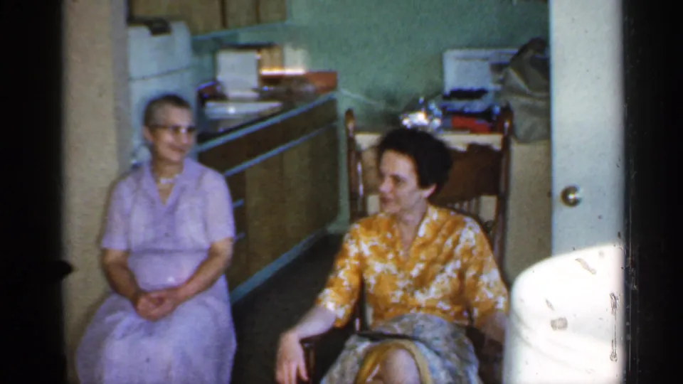 Two women remain seated in a 1959 Indiana private home interior, with kitchen cabinets and a doorway visible behind them.