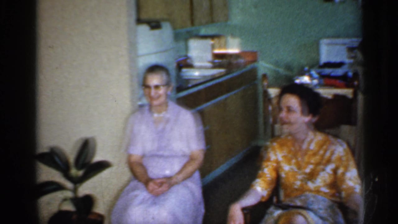Two women seated near a kitchen, one in lavender with glasses and one in a yellow patterned blouse.