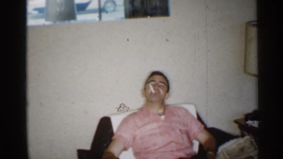 1959 Indiana home-movie frame showing a man in a pink shirt seated back in a chair while smoking indoors.