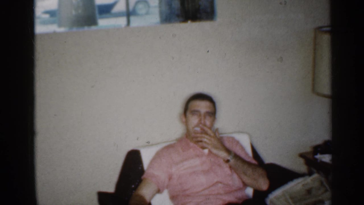 Man in a pink shirt sits in a living room and smokes a cigarette.