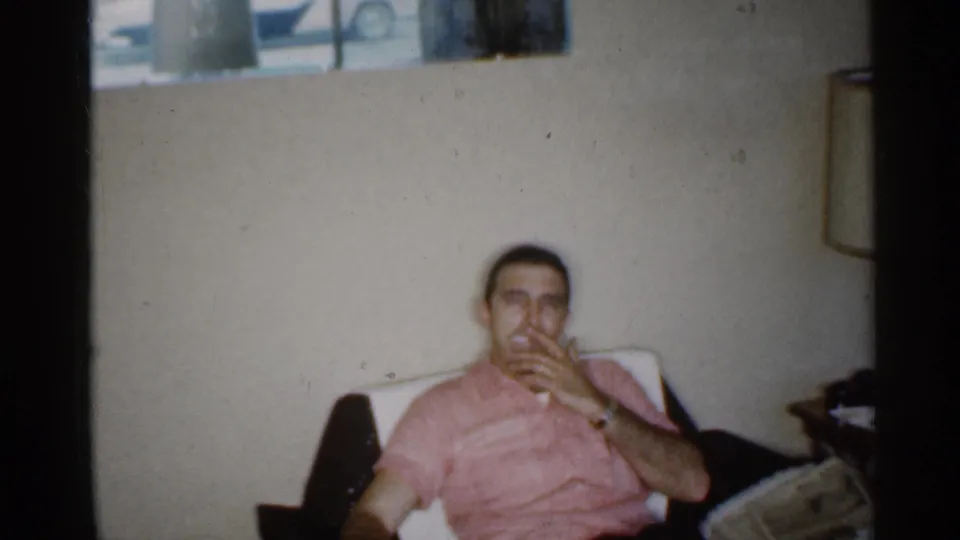 In 1959 Indiana home-movie footage, a man in a pink shirt sits on upholstered seating in a private living room while smoking.