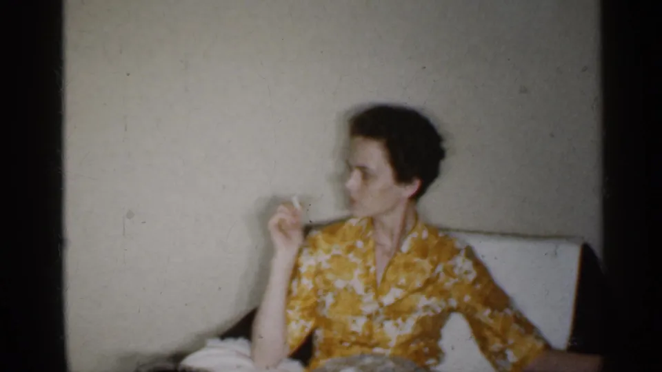 In 1959 Indiana home-movie footage, a woman in a yellow floral dress sits against a plain wall while holding a cigarette.