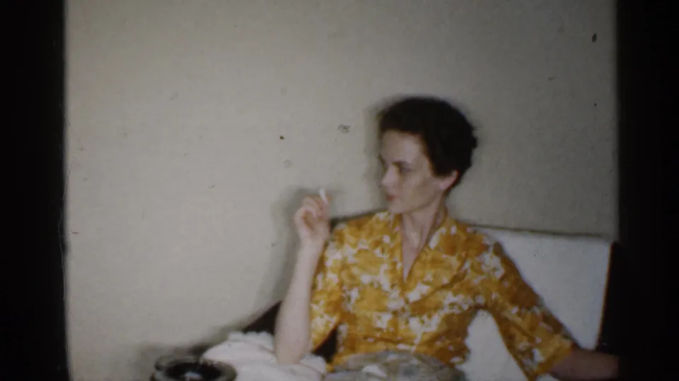 The woman remains seated in the private Indiana living room, holding a cigarette near her face in soft 8mm color footage.