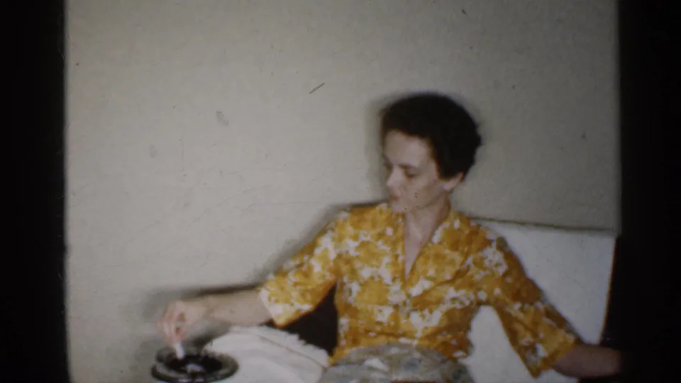 The woman in the yellow floral dress lowers her cigarette toward a dark ashtray on the seat or table beside her.