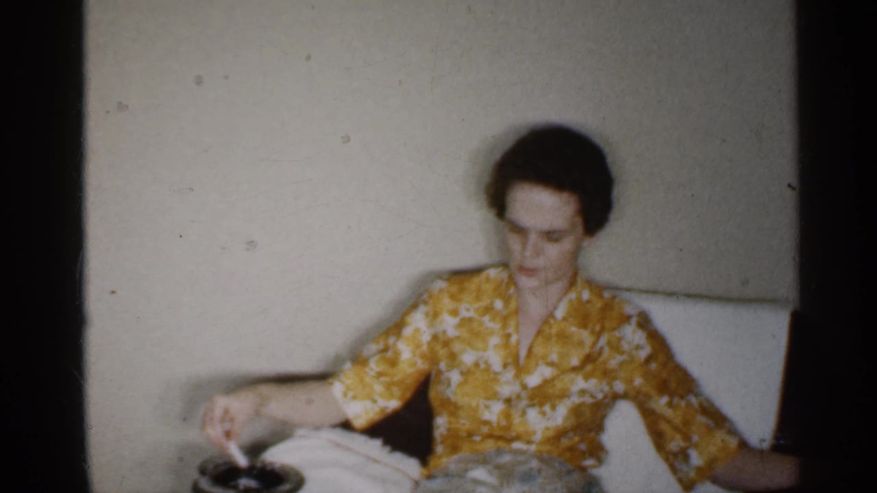 Woman in a yellow floral dress seated on a couch beside an ashtray.