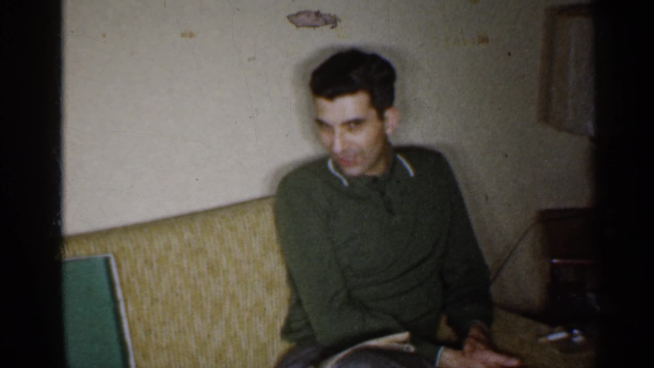 In a 1959 Indiana home-movie frame, a man in a green sweater sits on a couch near a side table and lamp.