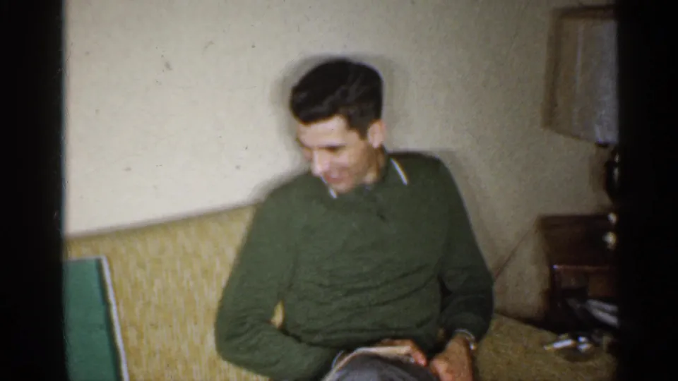 The 1959 Indiana home-movie frame shows a man in a green sweater looking down while seated on a couch in a living room.
