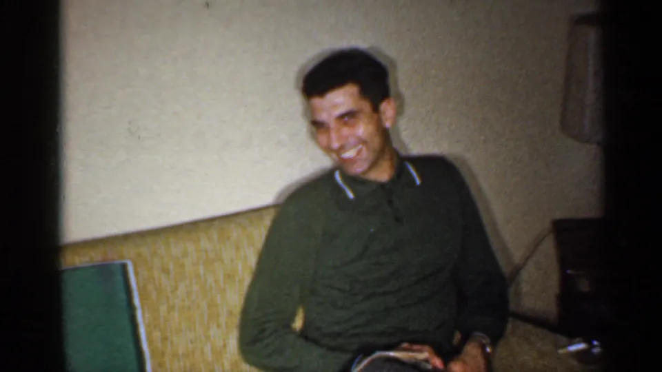 In a 1959 Indiana home-movie frame, a seated man in a green sweater smiles toward the camera from a living room couch.