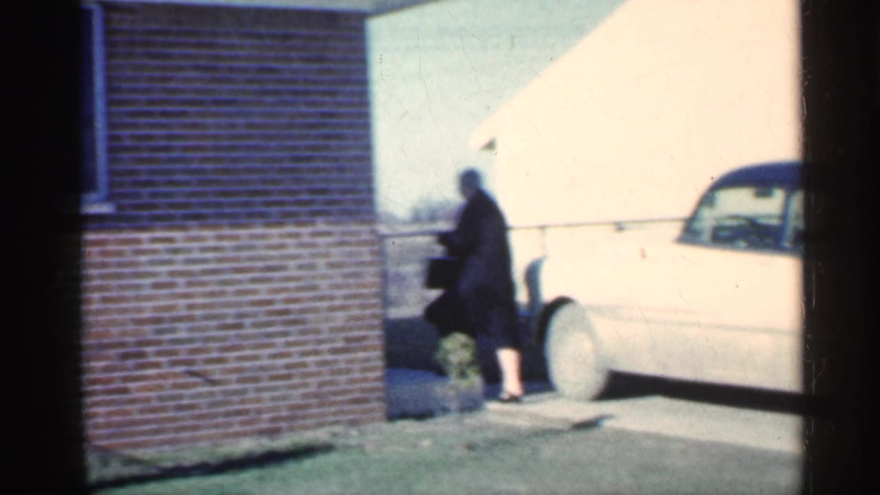 Woman in dark clothing walks beside a brick house near a light-colored vintage car.
