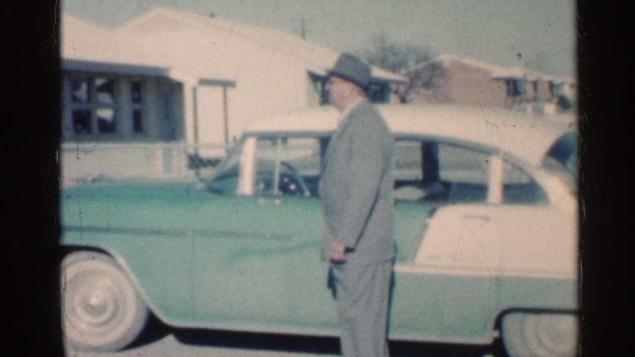 1959 Indiana frame of a man in a fedora beside a two-tone green and white mid-century car near a house.